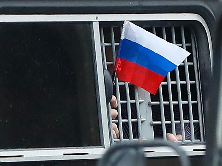 Ein festgenommener Demonstrant schwenkt aus einem Polizeiwagen heraus eine kleine russische Fahne.