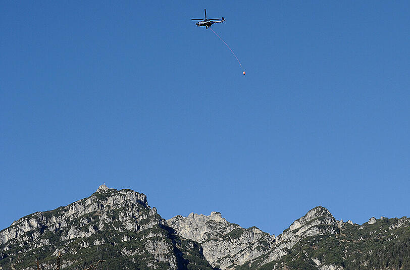 Ein Hubschrauber mit einer Löschbehälter fliegt bei Garmisch-Partenkirchen über dem Kramer (Gipfel links) zur Einsatzstelle.