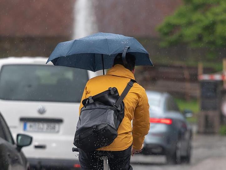 Ein Fahrradfahrer mit Regenschirm auf der Münchener Maximiliansbrücke. Foto: Peter Kneffel/Archivbild