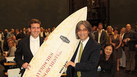 Florian Gmelin, Kontrabassist im Bayerischen Staatsorchester, überreicht Kent Nagano ein von den Musikern signiertes Surfboard. Rechts Naganos Gattin, die Pianistin Mari Kodama.