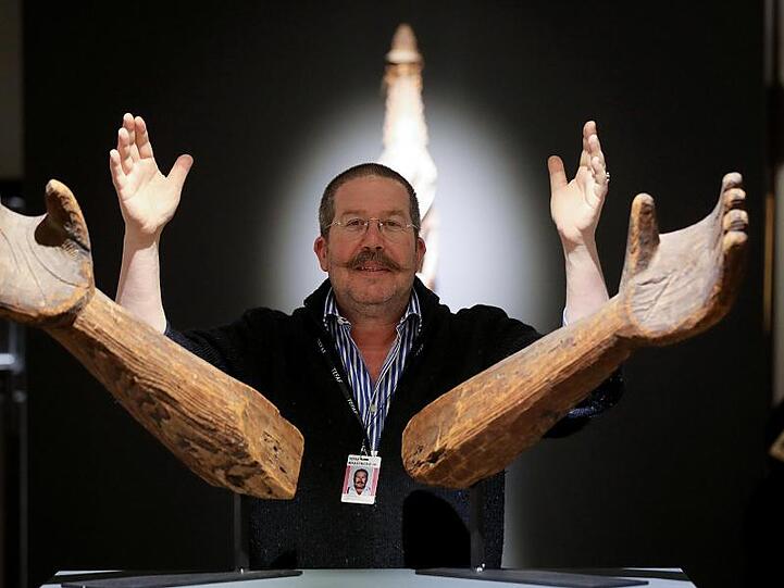 Galerist Anthony Meyer auf dem Stand seiner Galerie auf der Kunstmesse TEFAF hinter Unterarmen aus Holz eines Indianerstammes aus British Columbia.