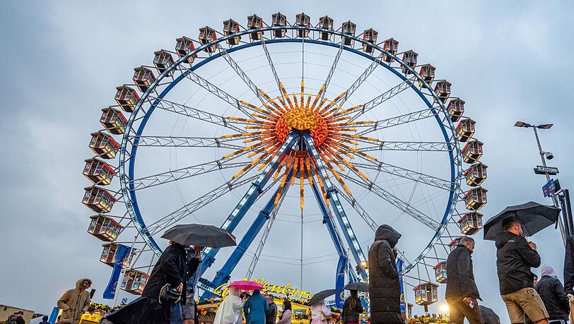 Besucher des Oktoberfestes in München gehen im Regen über das Festgelände vor dem Riesenrad.
