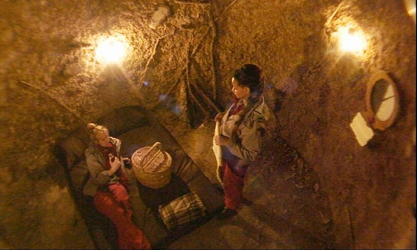 In einer Erdhöhle befindet sich eine Pritsche, Essen und Wein. An der Wand hängt eine Uhr, bei der eine Seite abgeklebt ist.
