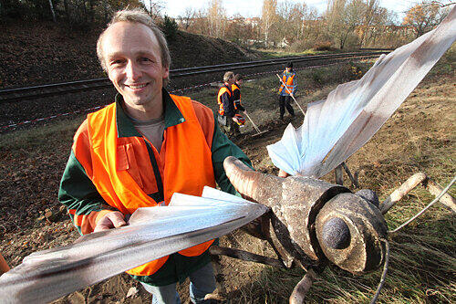 Landschaftspfleger Petr Mlnarik mit einem Modell der Blauflügeligen Ödlandschrecke. Hinten harken Helfer die Böschung.