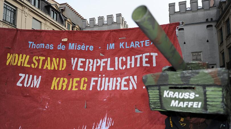 Ein Demonstrant mit Panzerkostüm steht vor einem Transparent mit der Aufschrift "Thomas de Misere - Im Klartext - Wohlstand verpflichtet zum Kriegführen".