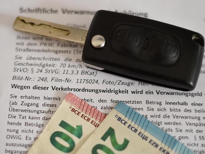 Eine schriftliche Verwarnung wegen zu schnellem Fahren, ein Autoschlüssel und Geldscheine. Foto: picture alliance / Angelika Warmuth/dpa