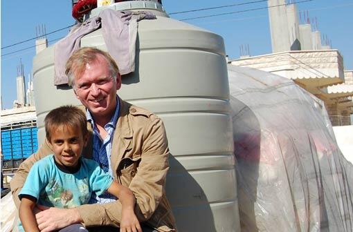 Der Münchner Kabarettist Christian Springer mit einem syrischen Flüchtlingskind vor einem Wassertank, den die Orienthelfer mit Spendengeldern errichtet haben.