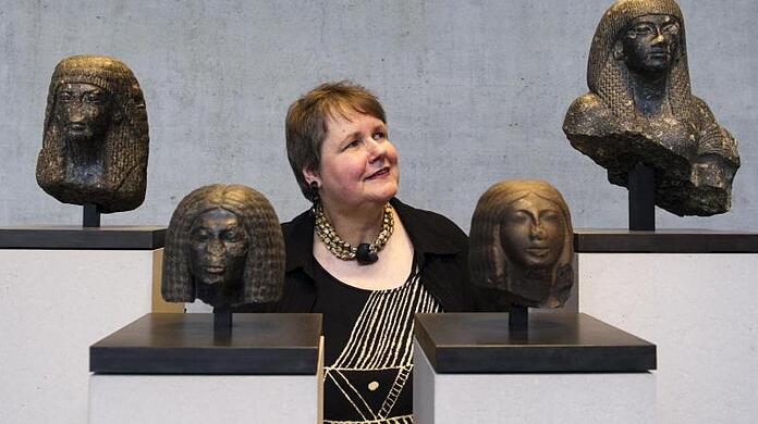 Sylvia Schoske steht zwischen vier Portraitköpfen.