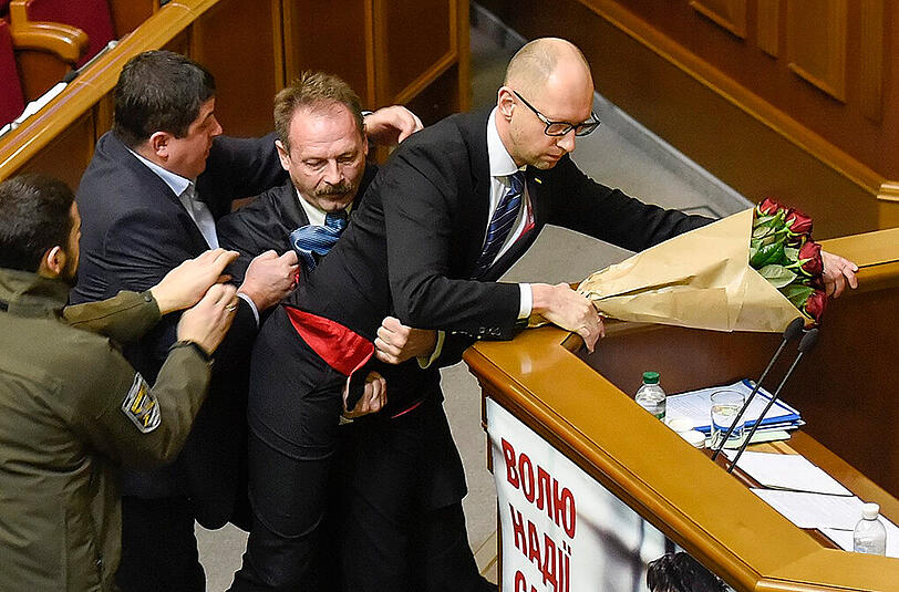Jazenjuk klammert sich am Rednerpult fest, während ihm ein Parlamentarier in den Schritt greift.