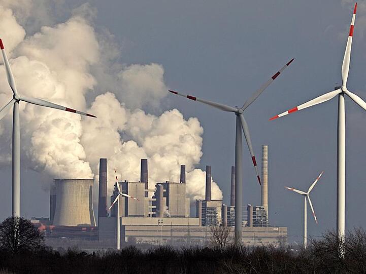 Alte und neue Technik: Rauch und Dampf steigen aus einem Braunkohlekraftwerk, im Vordergrund stehen Windräder.