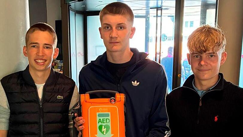 Drei junge Allg&auml;uer reanimieren in M&uuml;nchen einen Mann auf offener Stra&szlig;e und halten im Anschluss stolz ein lebensrettendes Hilfsmittel in die Kamera: den Automatisierten externen Defibrillator.