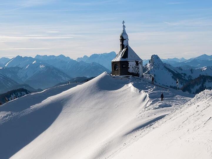 Skifahrer laufen zur Piste unterhalb der vom Schnee bedeckte Wallberg-Kapelle "Heilig Kreuz". Foto: Peter Kneffel