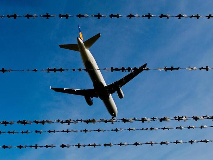 Ein Flugzeug fliegt hinter Stacheldraht am Himmel vorbei. Foto: Julian Stratenschulte/dpa/Symbolbild