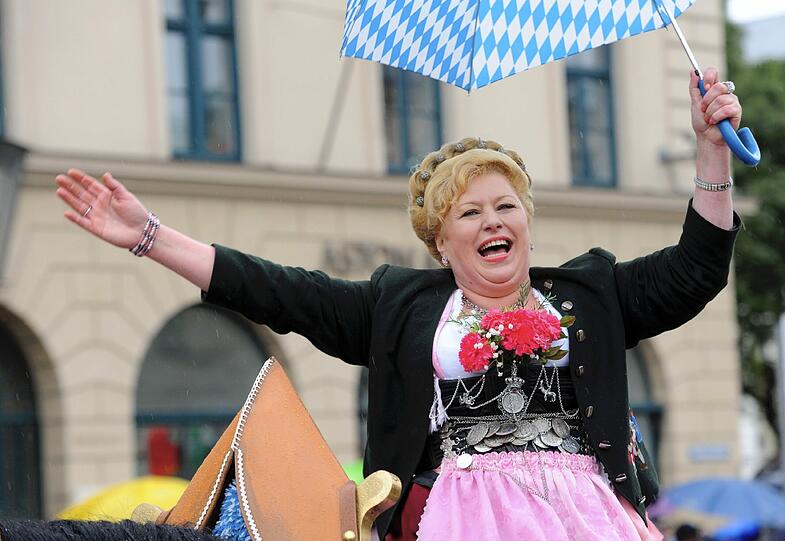 Der blau-weiße Regenschirm könnte fast zur Tracht dazugehören. Eine Dame winkt von ihrem Wagen aus in die Menge. Und ein zünftiger Zuschauer...