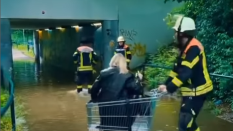 Feuerwehrleute helfen Passanten mit einem Einkaufswagen über eine überschwemmte Unterführung.