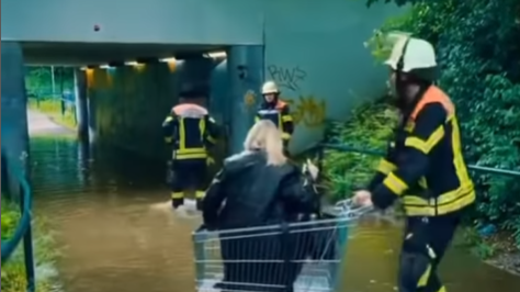 Feuerwehrleute helfen Passanten mit einem Einkaufswagen über eine überschwemmte Unterführung.