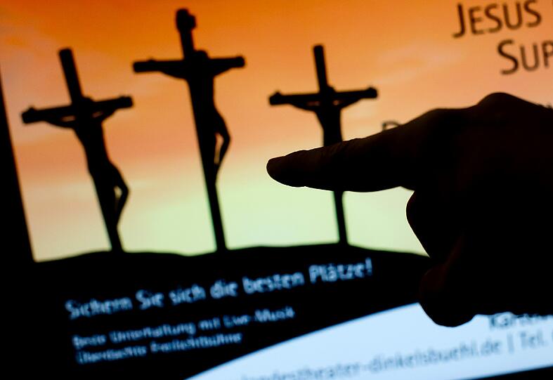 Ein Mann zeigt am 04.02.2015 in München (Bayern) auf ein Plakat des Landestheaters Dinkelsbühl, welches auf einem Computermonitor dargestellt wird. Das Plakat sorgt für große Aufregung. Es zeigt im Schattenriss drei gekreuzigte Menschen und darunter steht: «Sichern Sie sich die besten Plätze!» .
