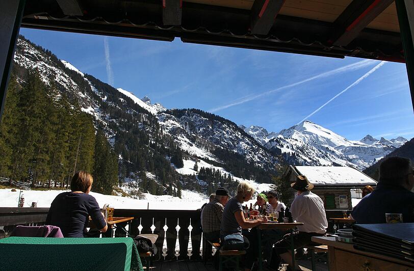 Aber nicht nur Frühlings- und Sommerfans freuen sich über das Wetter. Ausflügler sitzen auf der Terrasse eines Gasthauses im Stillachtal bei Oberstdorf im Sonnenschein vor dem Panorama der schneebedeckten Alpen.