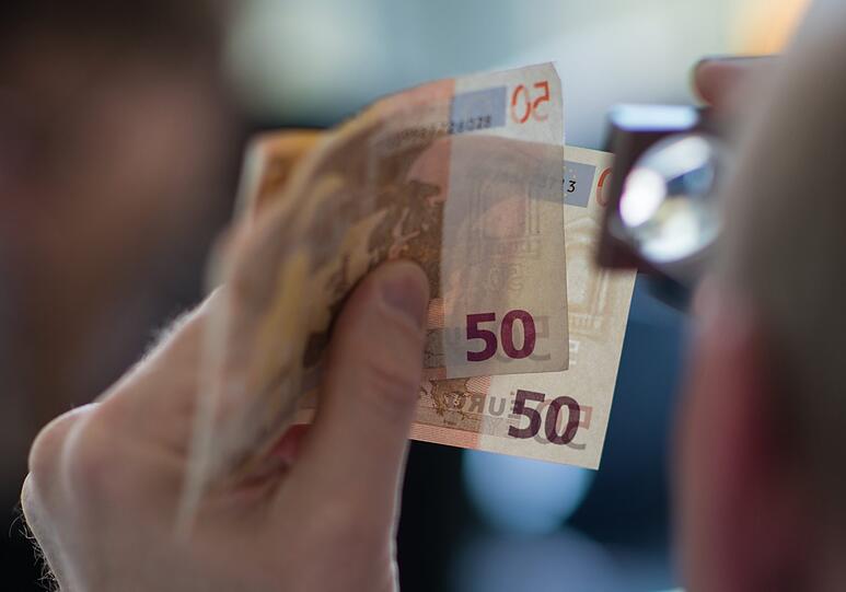 Ein Mann prüft mit einer Lupe zwei 50 Euro-Banknoten.