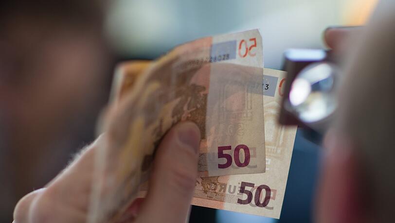 Ein Mann prüft mit einer Lupe zwei 50 Euro-Banknoten.