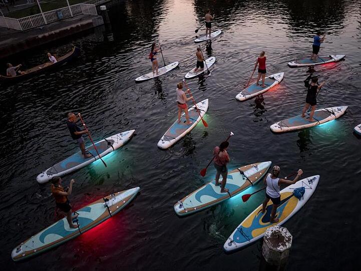 Teilnehmer einer Ausflugsgruppe fahren mit leuchtenden LED-Stand-Up-Paddles in den Abendstunden über einen Alsterfleet in Hamburg.