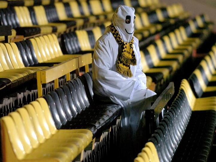 Ein als Geist verkleideter Mann sitzt auf den leeren Rängen eines Stadions. Foto: Oliver Berg/dpa/Symbolbild