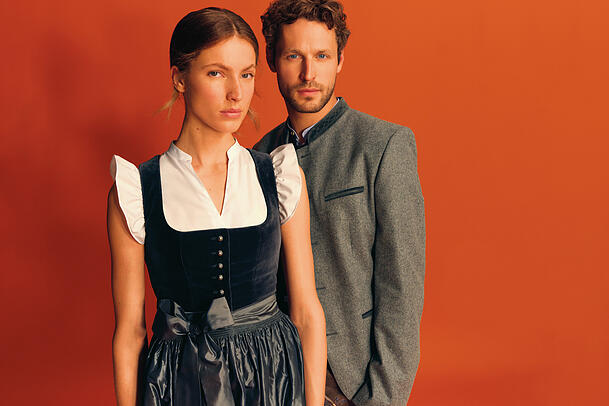 Sie in einem graublauen Dirndl mit Samtmieder und Seidenschürze, er im grauen Lodenjanker aus Schurwolle – beide Modelle von "Heimatwerk Ludwig Beck". Sie in einem graublauen Dirndl mit Samtmieder und Seidenschürze, er im grauen Lodenjanker aus Schurwolle – beide Modelle von "Heimatwerk Ludwig Beck".
