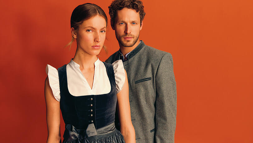 Sie in einem graublauen Dirndl mit Samtmieder und Seidenschürze, er im grauen Lodenjanker aus Schurwolle – beide Modelle von "Heimatwerk Ludwig Beck". Sie in einem graublauen Dirndl mit Samtmieder und Seidenschürze, er im grauen Lodenjanker aus Schurwolle – beide Modelle von "Heimatwerk Ludwig Beck".