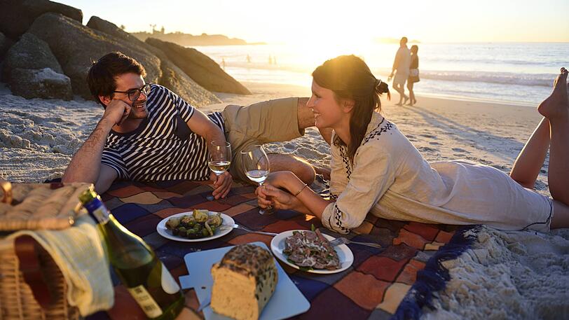 Ein junges Paar genießt ein Picknick am Strand. Mit dabei haben die beiden auch Brot und Alkohol. Aber im Urlaub darf man auch (mal) sündigen.