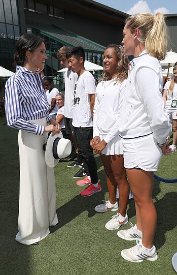 Herzogin Meghan begrüßt in weißer weit ausgestellter Hose und blau-weiß gestreifter Bluse Jugendspielerinnen in Wimbledon.