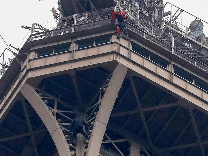 Ein Rettungshelfer (oben in roter Berufskleidung) klettert angeseilt am Eiffelturm hinunter, während unter ihm der Kletterer an die Gitterstäbe klammert.