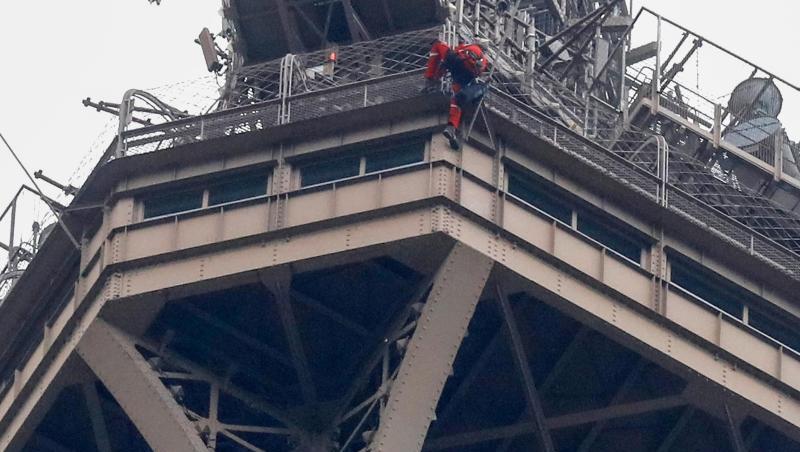 Ein Rettungshelfer (oben in roter Berufskleidung) klettert angeseilt am Eiffelturm hinunter, während unter ihm der Kletterer an die Gitterstäbe klammert. Ein Rettungshelfer (oben in roter Berufskleidung) klettert angeseilt am Eiffelturm hinunter, während unter ihm der Kletterer an die Gitterstäbe klammert.