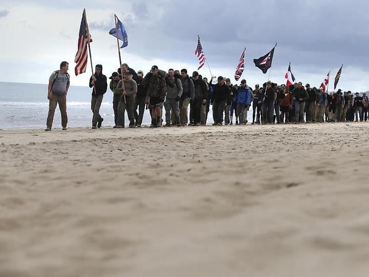 Männer gehen mit Flaggen der alliierten Truppen den Omaha-Beach in der Normandie entlang.