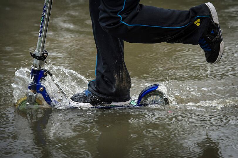 Ein anderes Kind flitzt mit dem Roller durch das Wasser.
