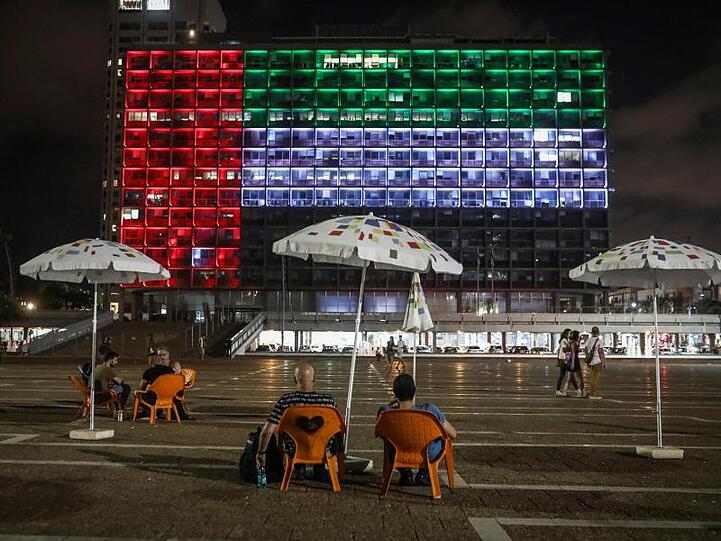 Menschen sitzen vor einem in den Nationalfarben der Emirate strahlenden Gebäude in Tel Aviv.