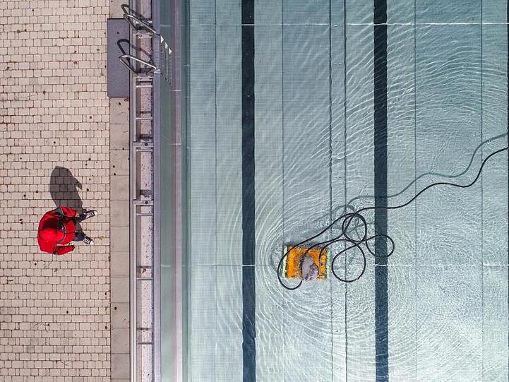 Ein Badleiter steht in einem Freibad am Beckenrand, und prüft die Arbeit eines Pool-Reinigungsroboters.
