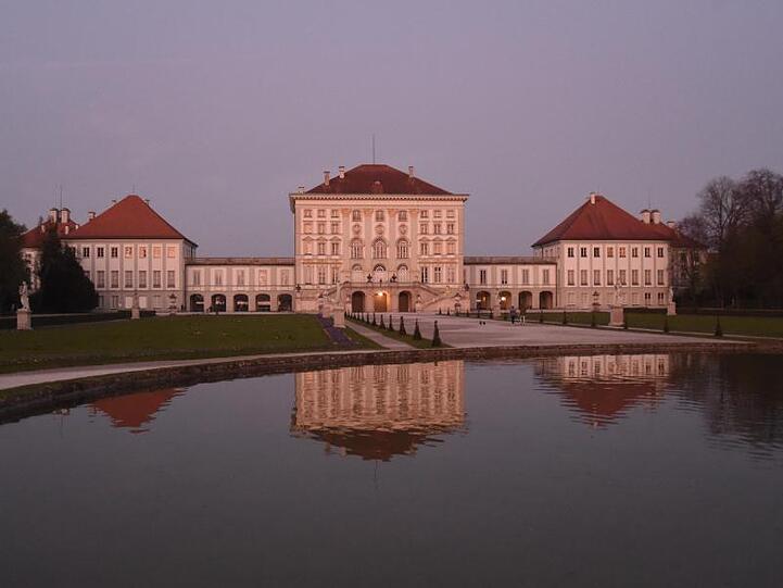 Das Schloss Nymphenburg wird von der Abendsonne rot beleuchtet und spiegelt sich im Wasser eines Brunnens. Foto: Felix Hörhager/dpa/Archivbild