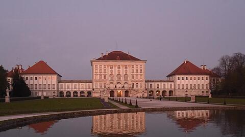 Das Schloss Nymphenburg wird von der Abendsonne rot beleuchtet und spiegelt sich im Wasser eines Brunnens. Foto: Felix Hörhager/dpa/Archivbild