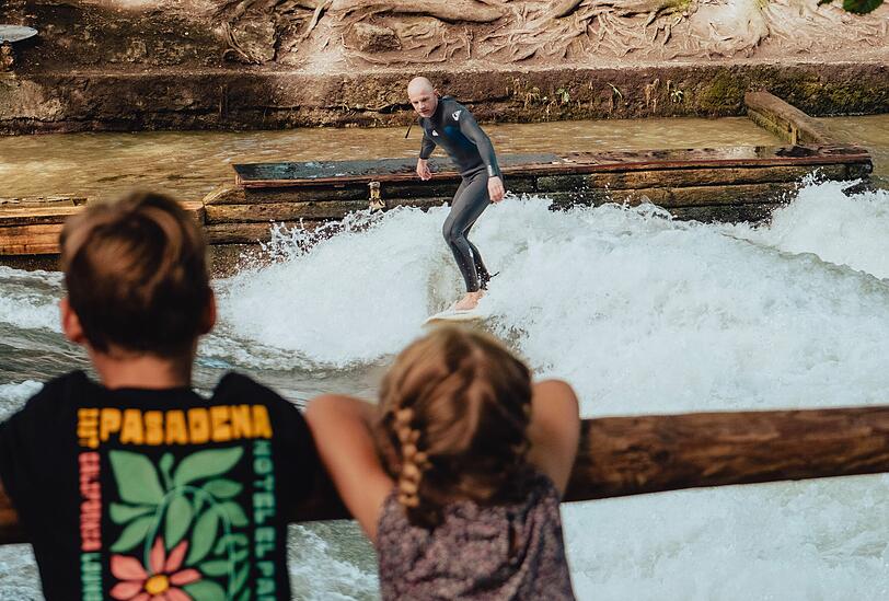 Zwei Kids schauen Zahnarzt Leopold beim Surfen zu. Ohnehin sammeln sich wieder sehr schnell Schaulustige, wenn sich jemand auf die schmale Welle wagt.
