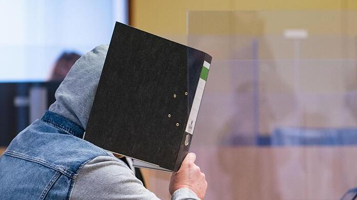 Der Angeklagte hält sich im Gerichtssaal einen Ordner vor das Gesicht.
