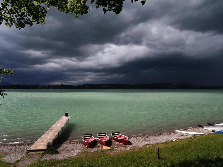 Eine Wolkenfront türmt sich hinter dem noch im Sonnenschein liegenden Forggensee an dessen Ufer ein Mann sitzt. Foto: Karl-Josef Hildenbrand