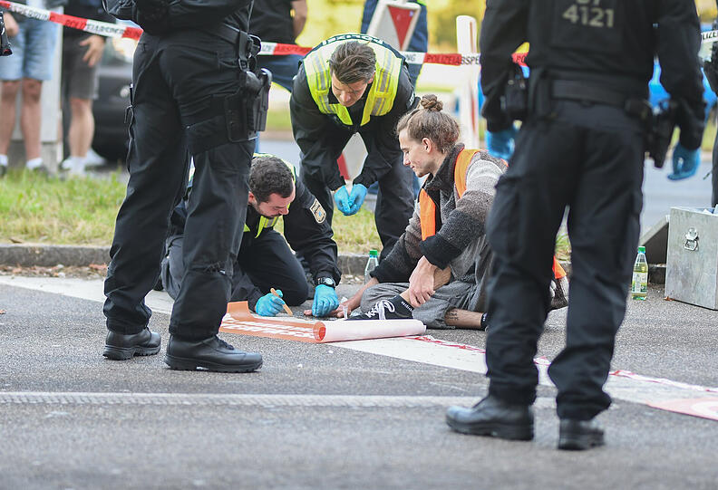 IAA-Protest der "Letzten Generation": Polizisten lösen die Hand eines Aktivisten vom Asphalt, der sich an der Autobahn A8 am Innsbrucker Ring festgeklebt hat.