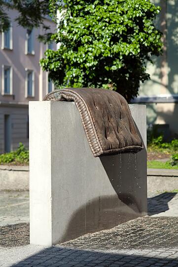 Tatiana Trouvé greift das im Stadtraum klassische Motiv des Brunnens auf. Ihre Skulptur kombiniert den Alltagsgegenstand einer Matratze aus hochwertigem Bronzeguss mit dem einfachen Baumaterial Beton.