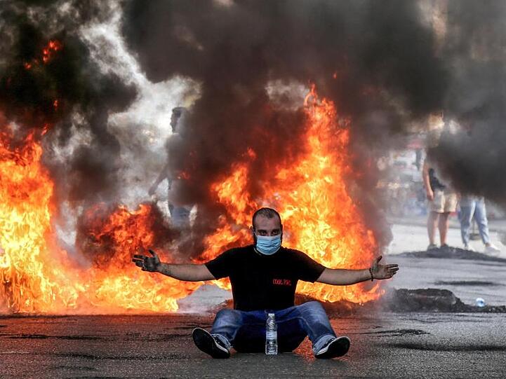 Ein Demonstrant mit Mundschutz sitzt mit ausgestreckten Armen vor einer brennenden Straßenbarrikade.