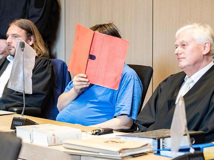 Der Angeklagte zu Beginn seines Prozesses mit einem Aktenmappe vor dem Gesicht im Landgericht im Gerichtssaal zwischen seinen Anwälten.