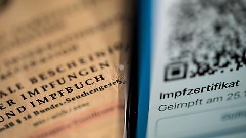 Ein Smartphone, auf dem ein Impfzertifikat angezeigt wird, liegt auf einem Impfbuch.