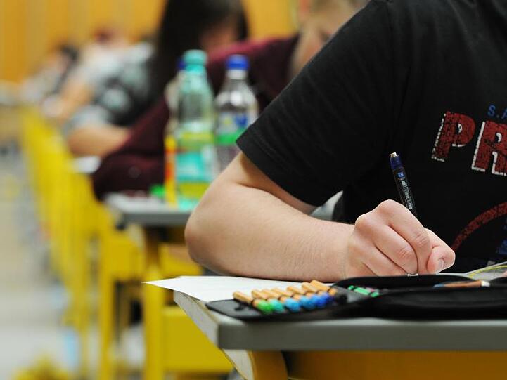 Schüler und Schülerinnen sitzen in einem Klassenraum und schreiben eine Prüfung. Die Person im Bildvordergrund hält einen Stift beim Schreiben in der rechten Hand. Foto: Armin Weigel/Archiv