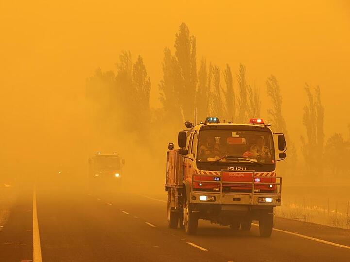 Feuerwehrfahrzeuge fahren in dichtem Rauch in der Region Canberra auf einer Straße, an deren Seiten das Gras brennt.