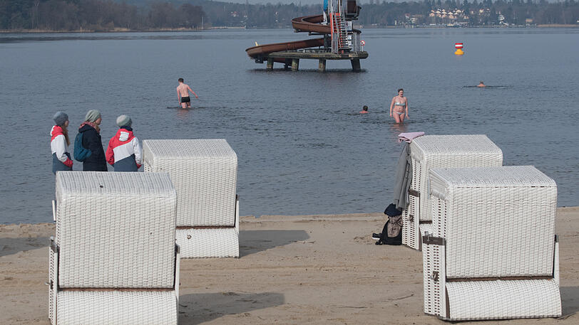 Drei Frauen stehen bei der Eröffnung der Freibädersaison im Strandbad Wannsee zwischen Strandkörben am Ufer und schauen den Schwimmern zu.