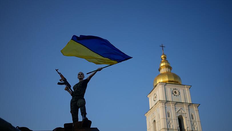 Eine Frau schwenkt die ukrainische Flagge auf einem zerstörten russischen Panzer in Kiew. Die Sonne scheint, aber es herrschen Traurigkeit und grimmige Entschlossenheit.